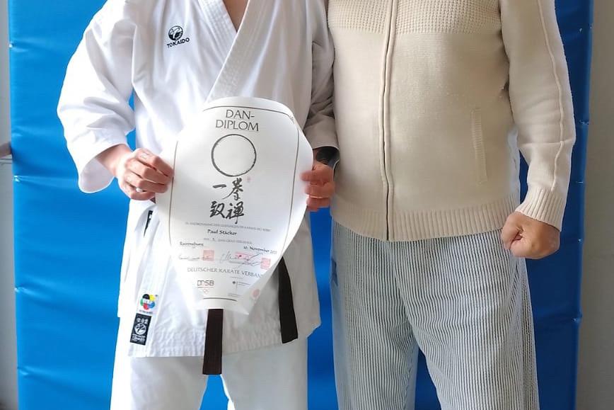 Karateka h&auml;lt sein Dan-Diplom und steht l&auml;chelnd neben einem &auml;lteren Mann vor einer blauen Mattenwand.