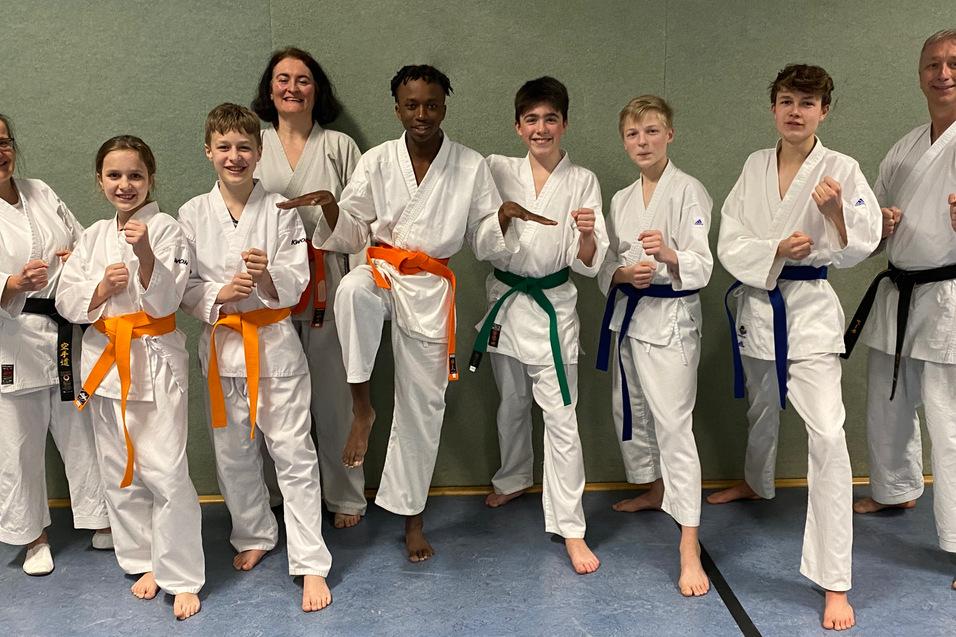 Gruppierung von acht Karateka in wei&szlig;en Gi, mit verschiedenen G&uuml;rtelfarben, posierend in einem Trainingsraum.