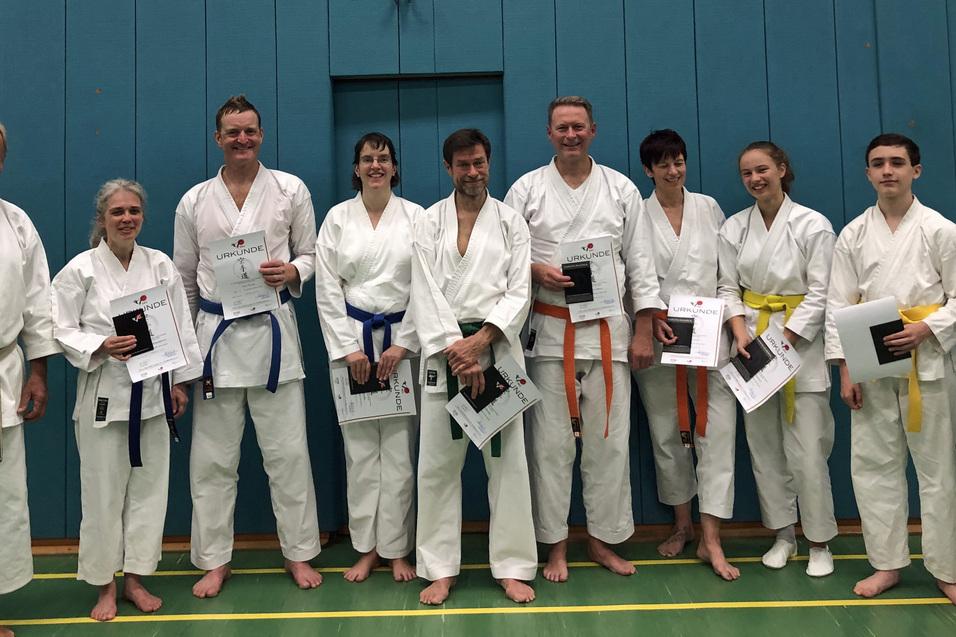 Gruppenfoto von Karatekas in wei&szlig;er Uniform, die stolz Urkunden in den H&auml;nden halten, vor einer blauen Wand.