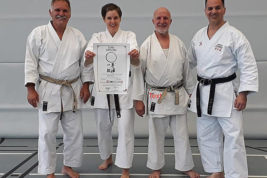Vier Karateka in wei&szlig;en Trainingsanz&uuml;gen posieren gemeinsam mit einem Dan-Diplom auf einer Matte.