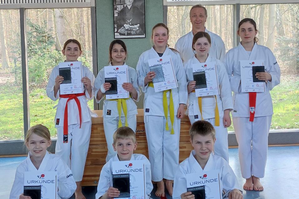 Eine Gruppe von Kindern in Karateanz&uuml;gen h&auml;lt Urkunden in der Hand, stehen vor einem Fenster mit Blick ins Freie.