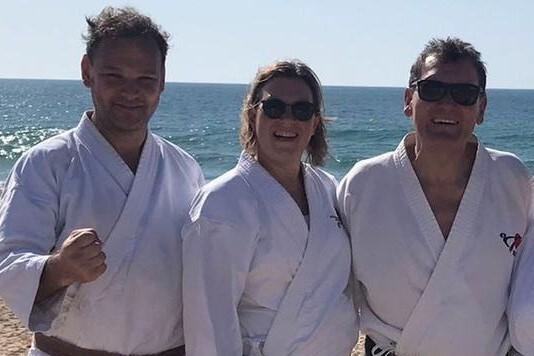 Drei Karateka in wei&szlig;en Gi mit Braungurt und zwei Schwarzgurten posieren l&auml;chelnd am Strand vor dem Meer.