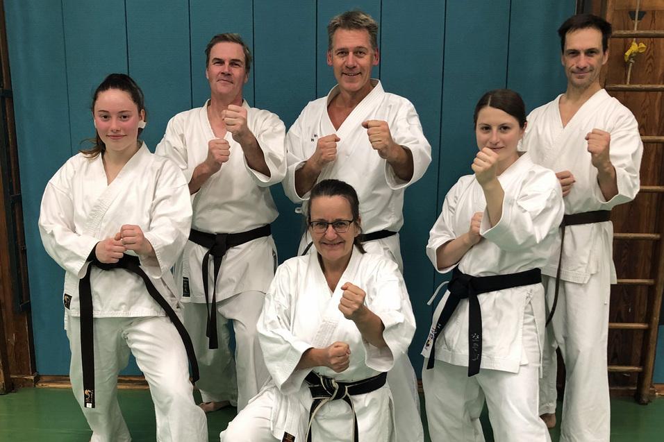 Sechs Karatek&auml;mpfer in wei&szlig;en Anz&uuml;gen und schwarzen G&uuml;rteln stehen grinsend in einer Kampfsporthalle.