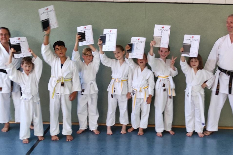 Prüfung 2023 Gruppe junger Karateka in Gi hält stolz Urkunden in einem Trainingsraum. Ein Trainer steht lächelnd daneben.