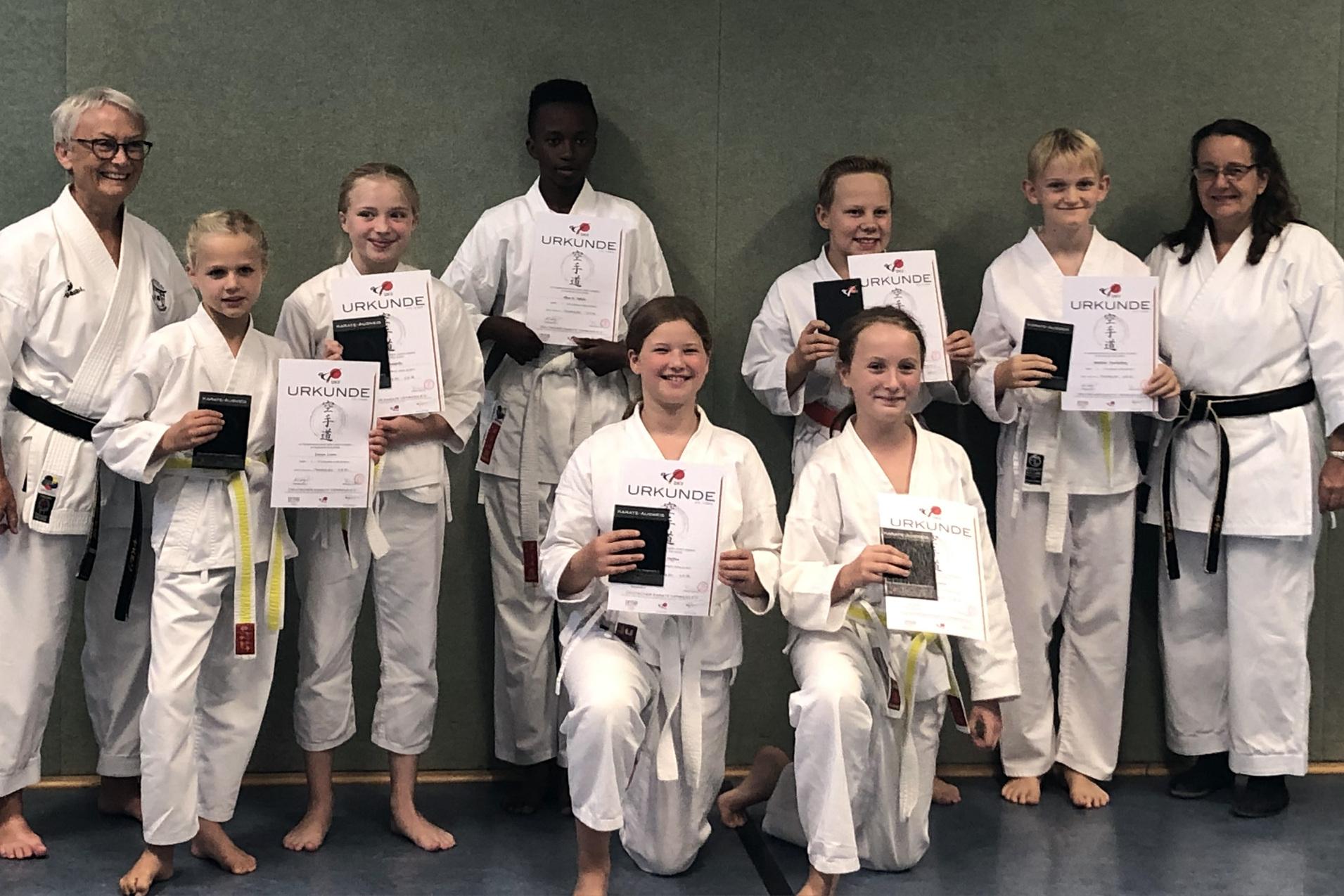 Gruppenfoto von Karateka in wei&szlig;em Gi, die Urkunden in den H&auml;nden halten, im Hintergrund eine gr&uuml;ne Wand.