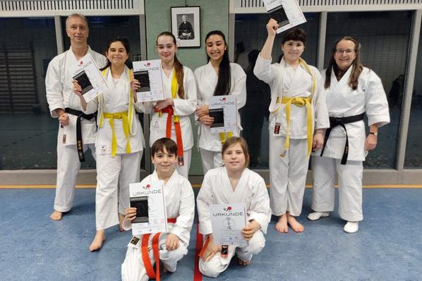 Gruppe von Karateka in wei&szlig;en Anz&uuml;gen h&auml;lt Urkunden in der Sporthalle, einen Lehrer mit schwarzem G&uuml;rtel im Hintergrund.