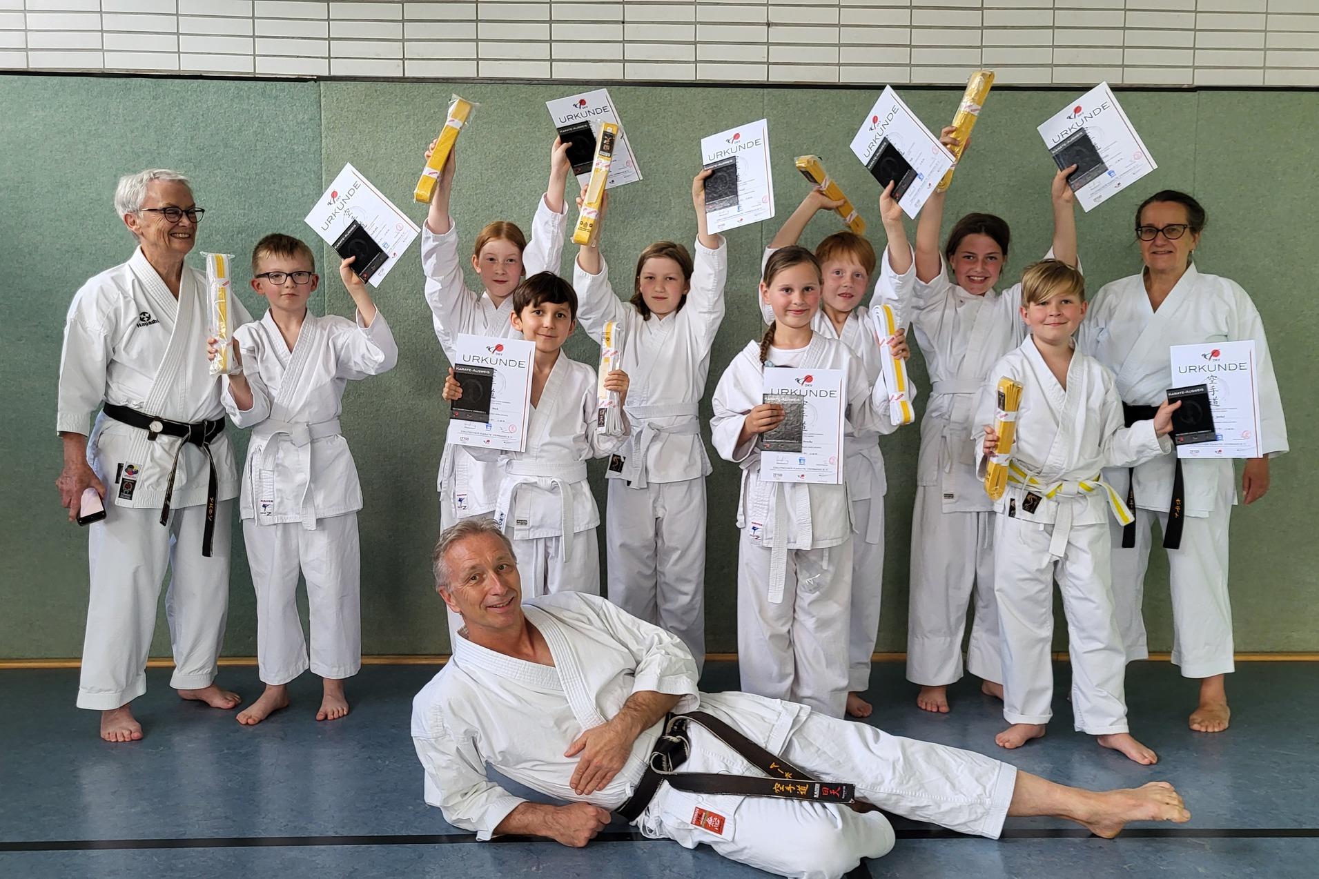 Gruppe von Karateka in Uniform h&auml;lt Urkunden und Medaillen, w&auml;hrend ein Trainer entspannt im Vordergrund posiert.