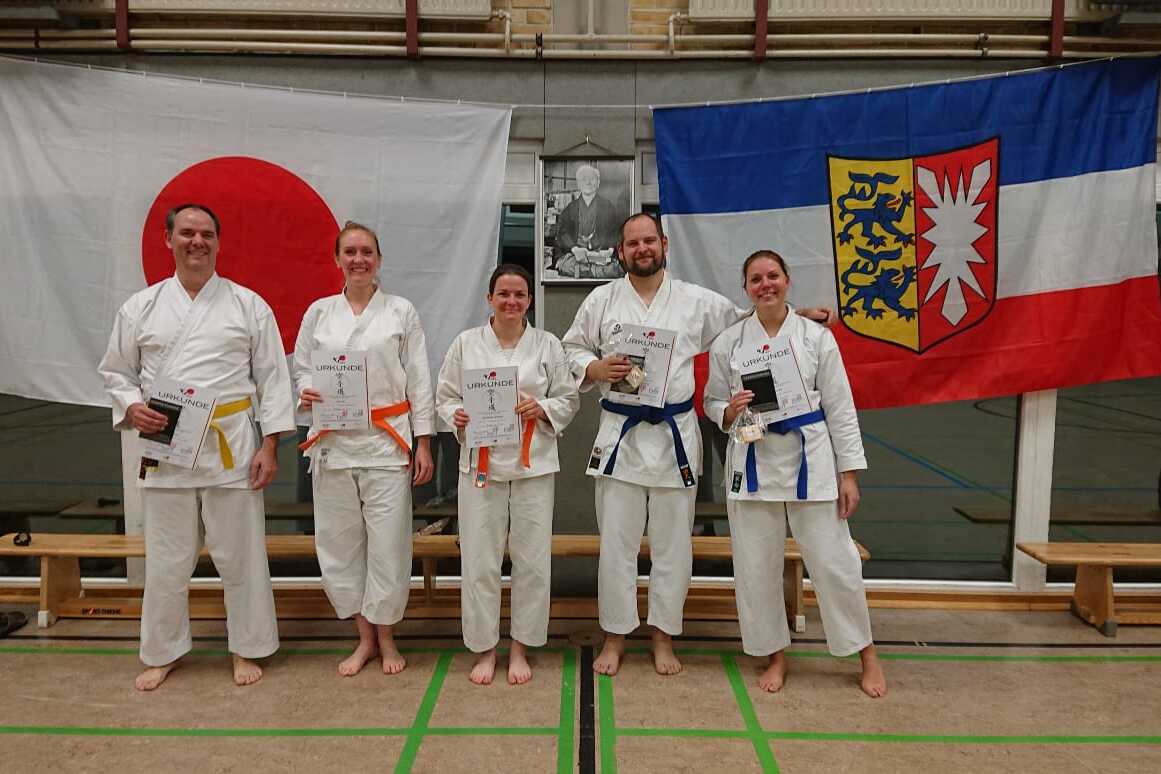 F&uuml;nf Karateka in wei&szlig;en Anz&uuml;gen halten Urkunden und stehen vor japanischen und deutschen Flaggen.