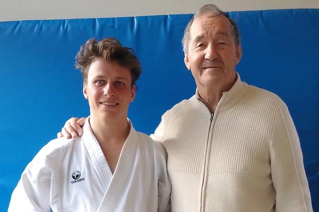 Karateka h&auml;lt sein Dan-Diplom und steht l&auml;chelnd neben einem &auml;lteren Mann vor einer blauen Mattenwand.