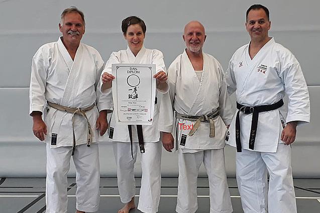 Vier Karateka in wei&szlig;en Trainingsanz&uuml;gen posieren gemeinsam mit einem Dan-Diplom auf einer Matte.