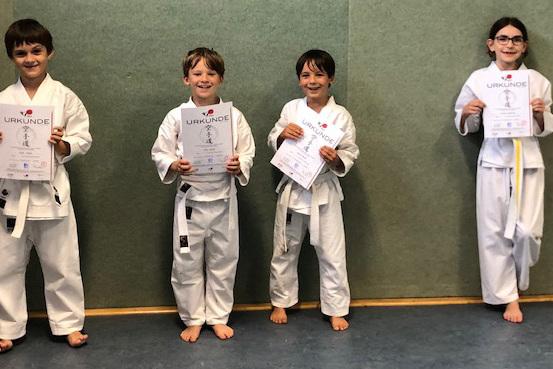 Vier Kinder in Karateanz&uuml;gen halten Urkunden in der Hand, stehend vor einer gr&uuml;nen Wand in einem Sportraum.