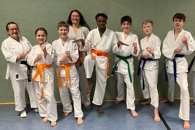 Gruppierung von acht Karateka in wei&szlig;en Gi, mit verschiedenen G&uuml;rtelfarben, posierend in einem Trainingsraum.