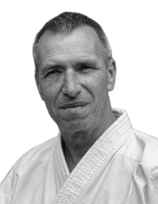 Porträt eines älteren Mannes in einem weißen Karate-Gi, lächelnd, vor schwarzem Hintergrund.