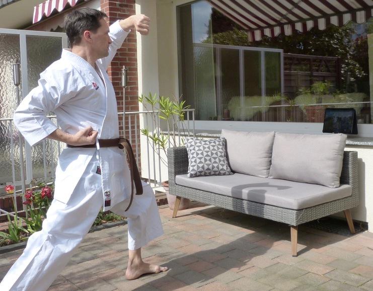 Mann in Karate-Kleidung führt eine Bewegung im Freien vor einer Couch mit Kissen und einem Tablet durch.