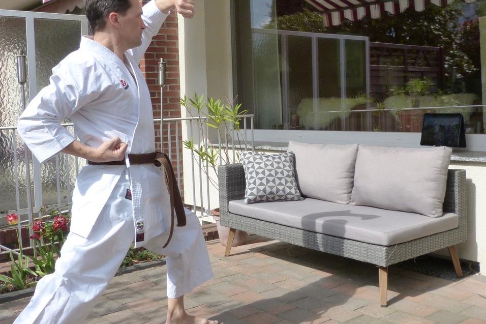 Mann in Karate-Kleidung f&uuml;hrt eine Bewegung im Freien vor einer Couch mit Kissen und einem Tablet durch.