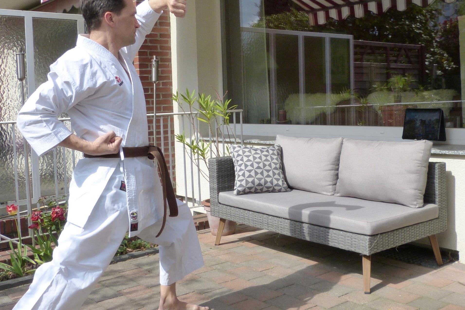 Mann in Karate-Kleidung führt eine Bewegung im Freien vor einer Couch mit Kissen und einem Tablet durch.