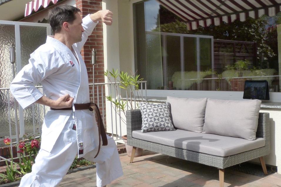 Mann in Karate-Kleidung f&uuml;hrt eine Bewegung im Freien vor einer Couch mit Kissen und einem Tablet durch.