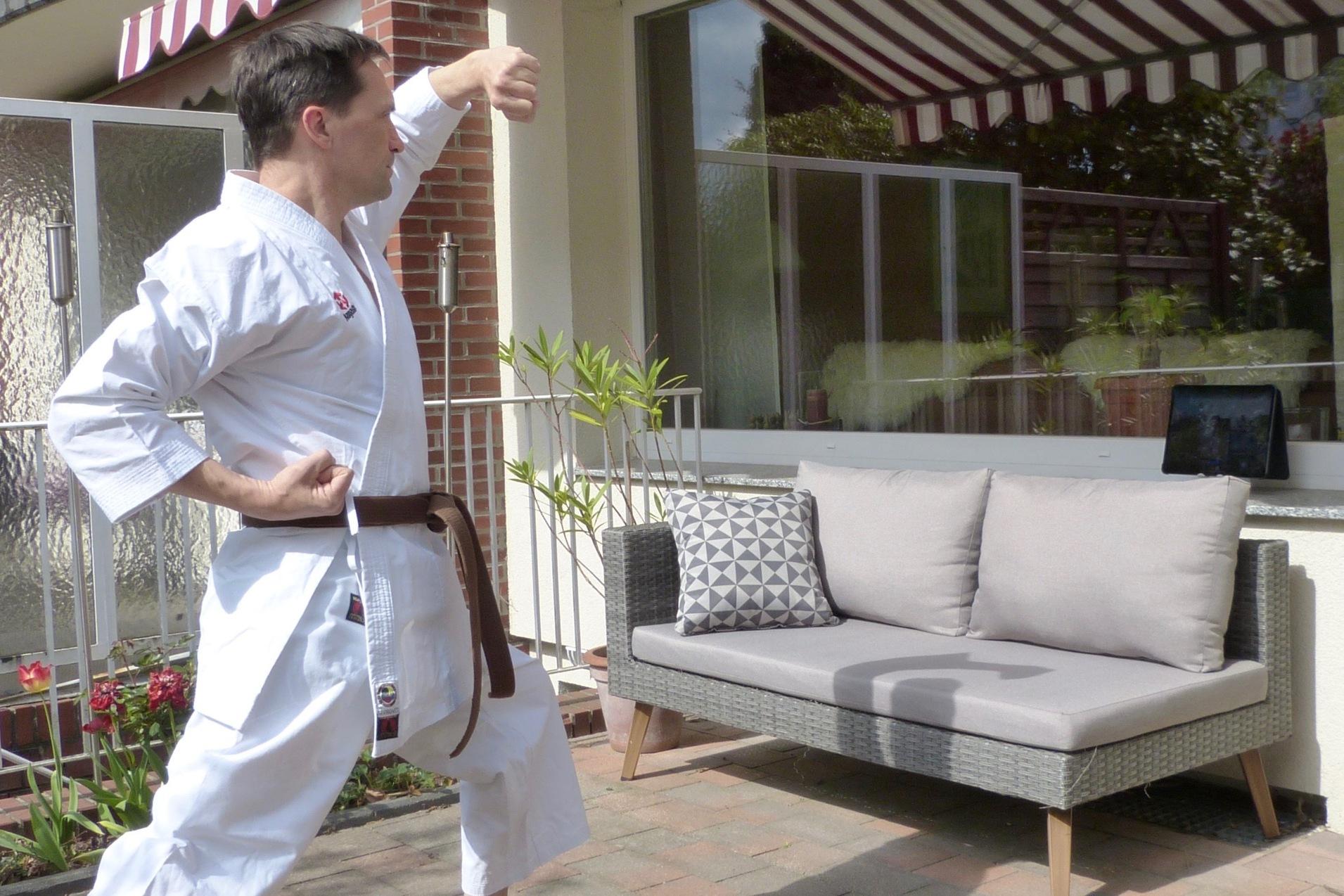 Mann in Karate-Kleidung f&uuml;hrt eine Bewegung im Freien vor einer Couch mit Kissen und einem Tablet durch.