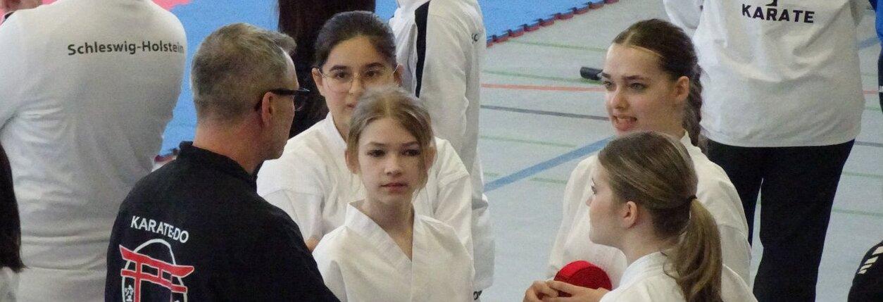 Mehrere Personen in Karateanz&uuml;gen stehen in einer Halle mit blau-rotem Boden, einige tragen Jacken mit Aufschriften wie 'Schleswig-Holstein' und 'KARATE D VFL-PINNEBERG'