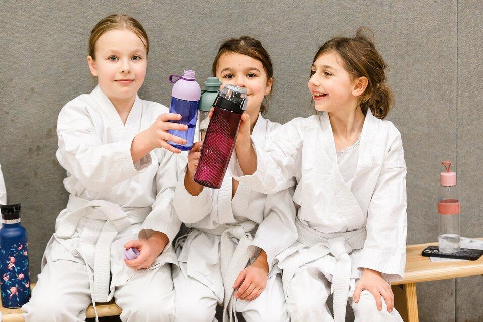 Drei Kinder in wei&szlig;en Karateanz&uuml;gen sitzen auf einer Bank und halten Trinkflaschen in die Kamera