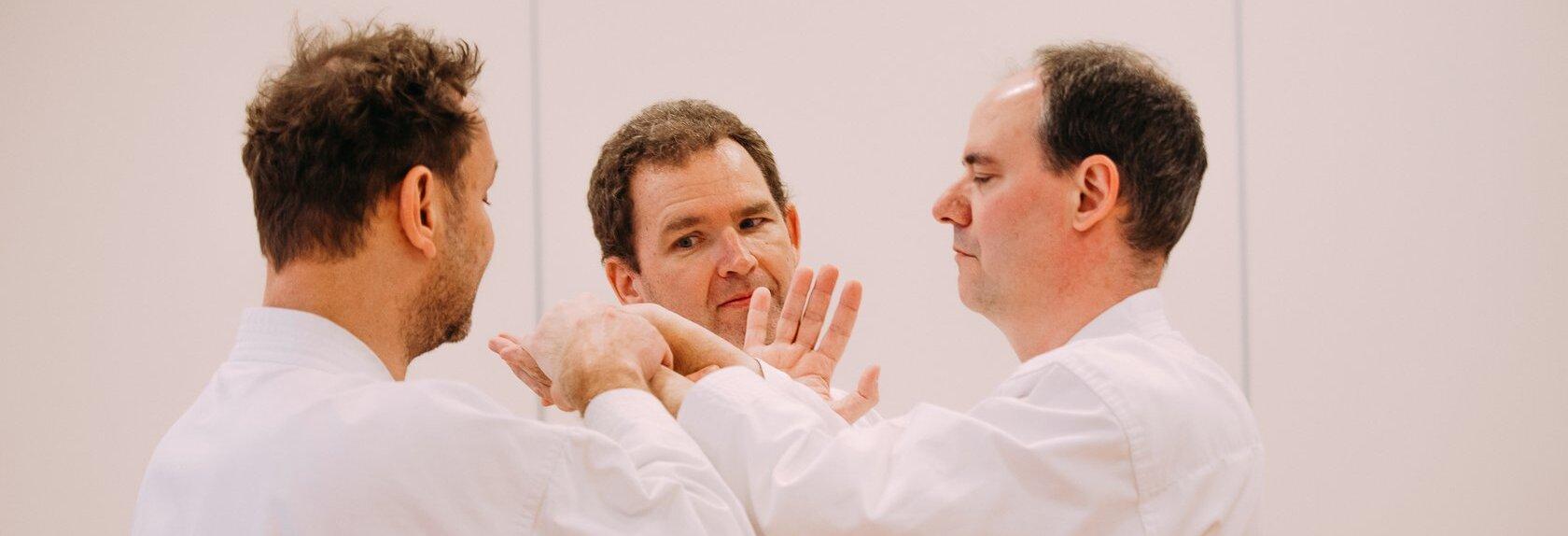 Drei Personen in wei&szlig;en Karateanz&uuml;gen stehen in einem Kreis und f&uuml;hren eine Handtechnik aus