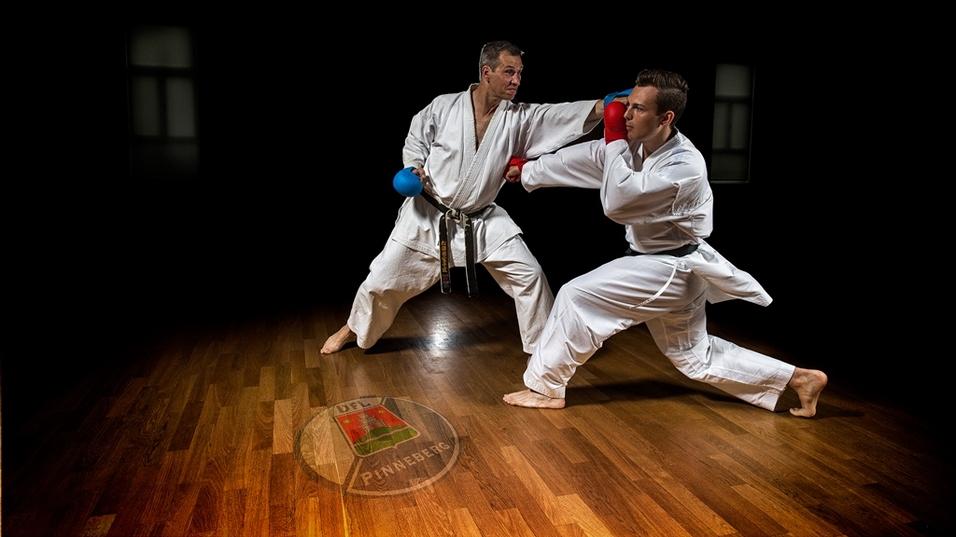 Zwei Karatekämpfer in weißen Anzügen üben Techniken auf einem Holzboden in einem dunklen Raum.