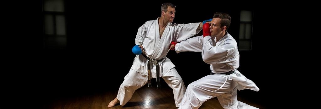 Zwei Karatek&auml;mpfer in wei&szlig;en Anz&uuml;gen &uuml;ben Techniken auf einem Holzboden in einem dunklen Raum.