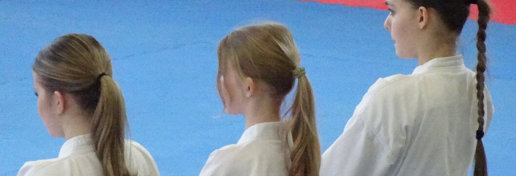 Drei Jugendliche in wei&szlig;en Karateanz&uuml;gen mit blauen G&uuml;rteln stehen in einer Reihe auf einer Matte mit rotem und blauem Bereich