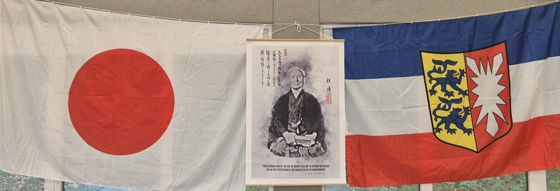 Drei Flaggen hängen an einer Wand: die japanische Flagge, eine historische Abbildung und eine Flagge eines deutschen Bundeslandes.