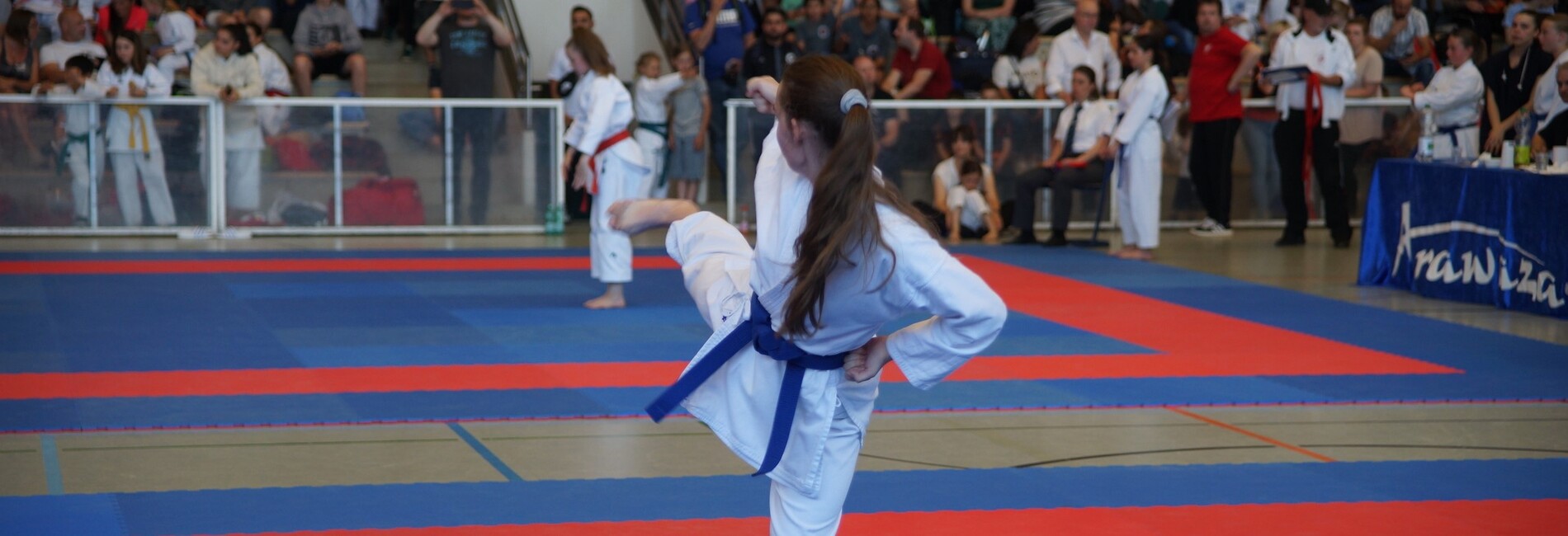 Ein Mädchen in Karate-Uniform führt einen Kick auf einer roten und blauen Matte bei einem Wettkampf aus.