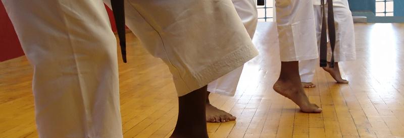 Vier Karateka in Kimonos stehen mit einer Beinposition auf einem Holzboden. Hintergründe in Blau und Rot sind sichtbar.