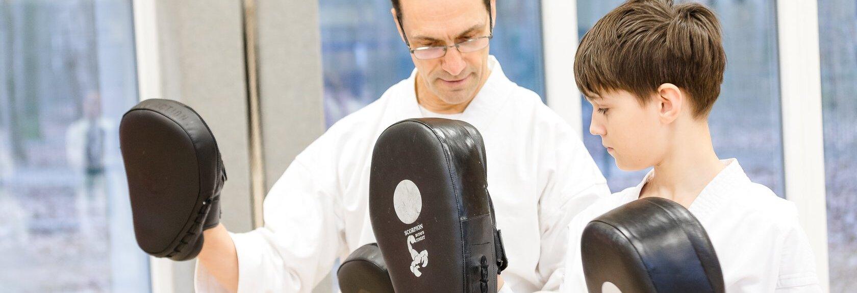 Zwei Personen in wei&szlig;en Karateanz&uuml;gen mit schwarzen und orangen G&uuml;rteln halten Schlagpolster in einer Trainingshalle