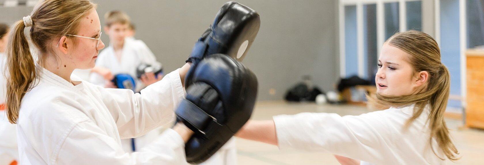 Zwei M&auml;dchen in Karateanz&uuml;gen mit farbigen G&uuml;rteln trainieren in einer Sporthalle, eines h&auml;lt Schlagpolster, das andere f&uuml;hrt einen Faustschlag aus