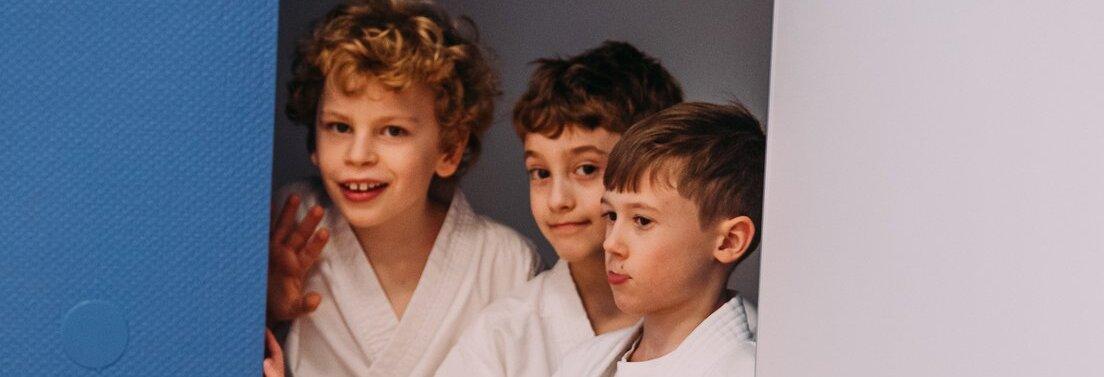 Drei Kinder in wei&szlig;en Karateanz&uuml;gen mit gelben G&uuml;rteln stehen nebeneinander an einer blauen und wei&szlig;en Wand