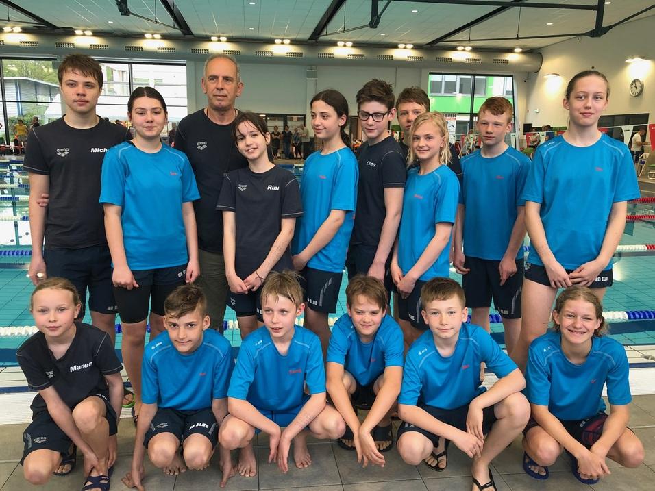 Gruppenshot von Schwimmern in blauen und schwarzen Trikots, stehend und kniend vor einem Schwimmbecken.