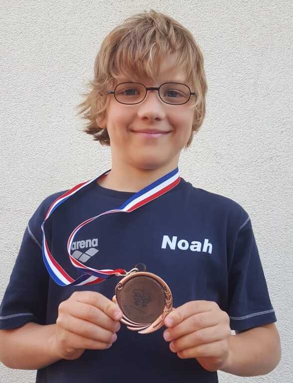 Junge mit Brille hält eine bronzene Medaille mit Aufdruck in der Hand, trägt ein blaues T-Shirt mit dem Namen „Noah“.