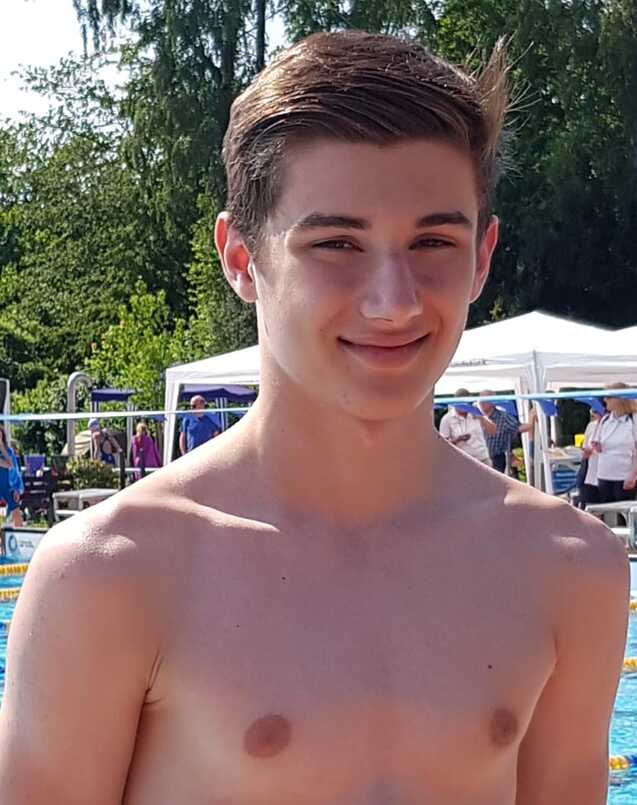 Junger Mann mit kurzem Haar steht lächelnd am Beckenrand eines Schwimmbads bei sonnigem Wetter.