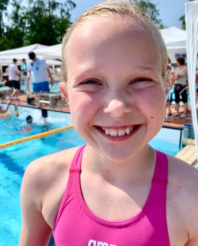 Ein lächelndes Mädchen mit kurzer blonder Frisur trägt einen pinken Badeanzug, Hintergrund zeigt ein Schwimmbecken.