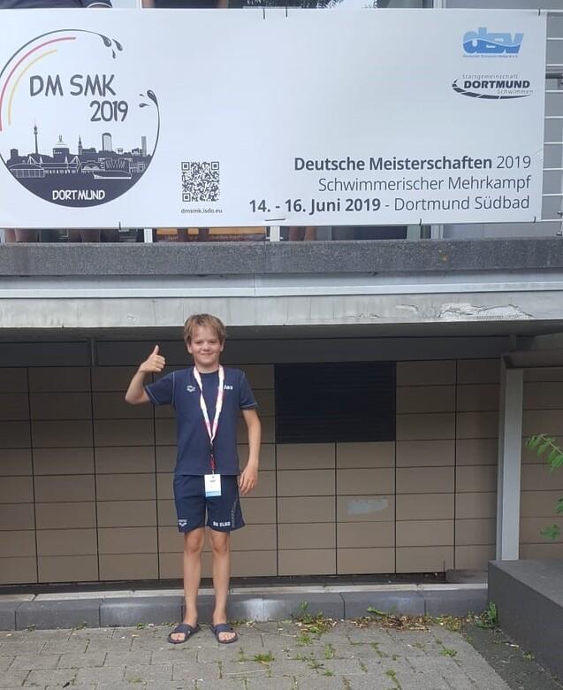 Junge mit Daumen hoch steht vor einem Plakat der Deutschen Meisterschaften im Schwimmmehrkampf 2019 in Dortmund.