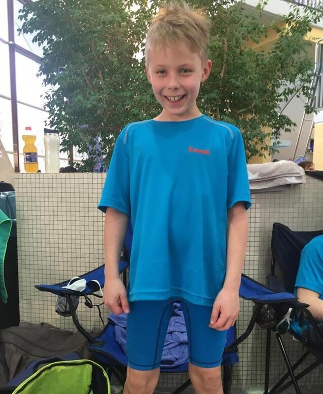 Lächelnder Junge in blauem Schwimmanzug steht vor einem Schwimmbad mit Pflanzen im Hintergrund.