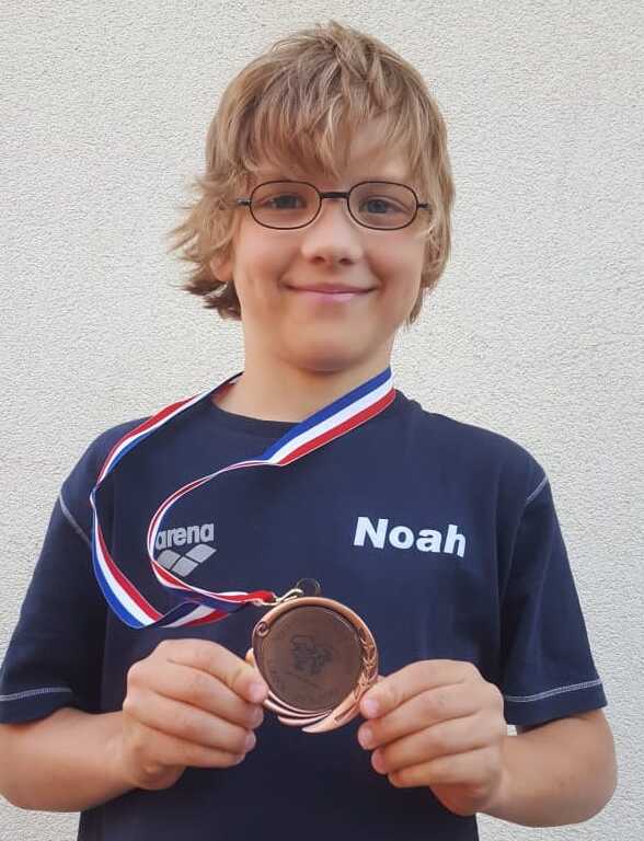 Junge mit Brille h&auml;lt eine bronzene Medaille mit Aufdruck in der Hand, tr&auml;gt ein blaues T-Shirt mit dem Namen &bdquo;Noah&ldquo;.