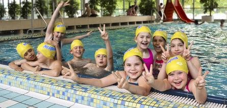 Gruppenkinder mit gelben Schwimmhauben posieren am Rand eines Schwimmbeckens und zeigen das Peace-Zeichen.