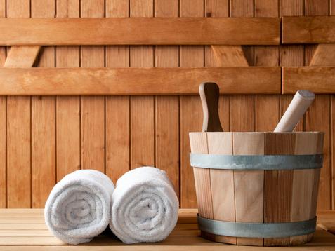 Handtücher und ein Holzgefäß mit einem Löffel stehen auf einer Holzliege in einer Sauna. Hintergrund aus Holzpaneelen.