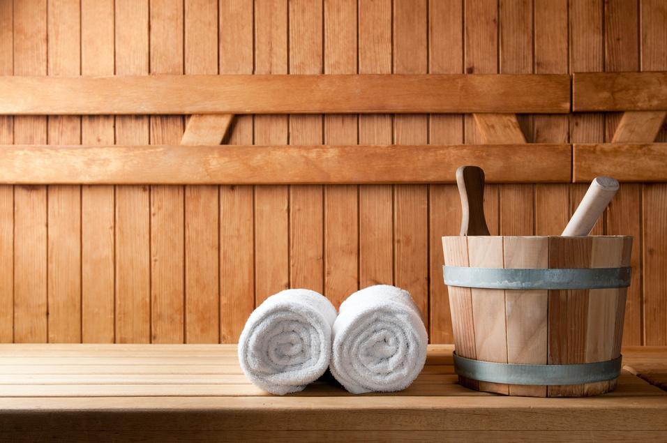 Handtücher und ein Holzgefäß mit einem Löffel stehen auf einer Holzliege in einer Sauna. Hintergrund aus Holzpaneelen.
