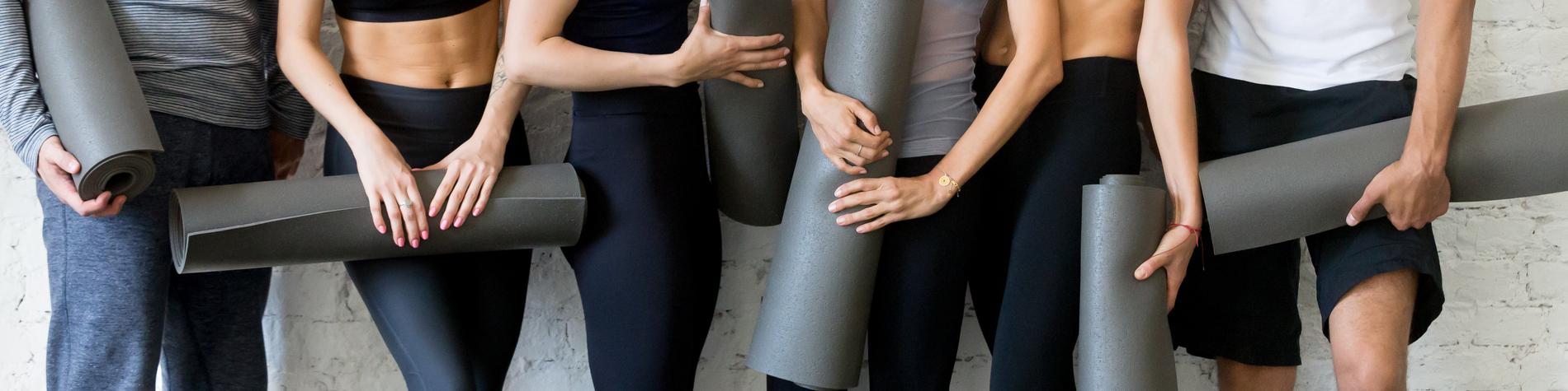 Sechs Personen in Sportkleidung lachen miteinander und halten Yogamatten in einem hellen Raum mit weißen Wänden.