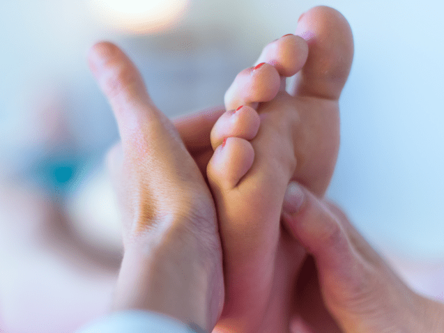 Nahaufnahme einer Hand, die eine Fußmassage an einem Fuß mit lackierten Nägeln durchführt. Hintergrund unscharf.