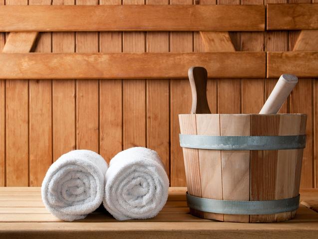 Handtücher und ein Holzgefäß mit einem Löffel stehen auf einer Holzliege in einer Sauna. Hintergrund aus Holzpaneelen.