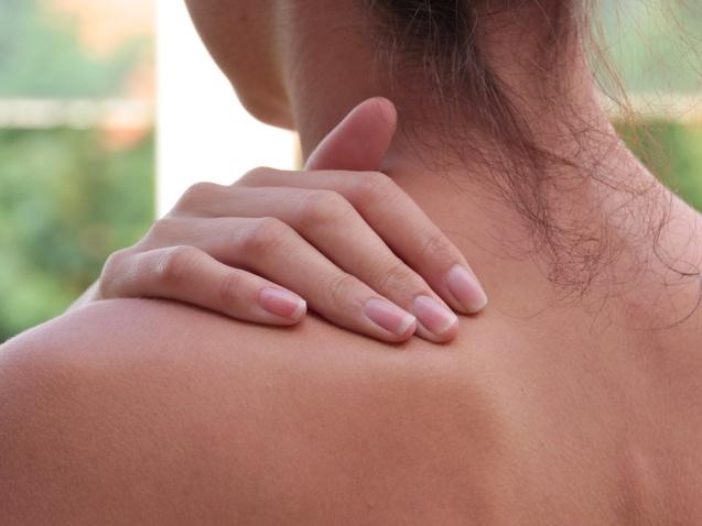 Nahaufnahme einer Frau, die sanft ihre Schulter mit einer Hand berührt, im Hintergrund unscharfe grüne Pflanzen.