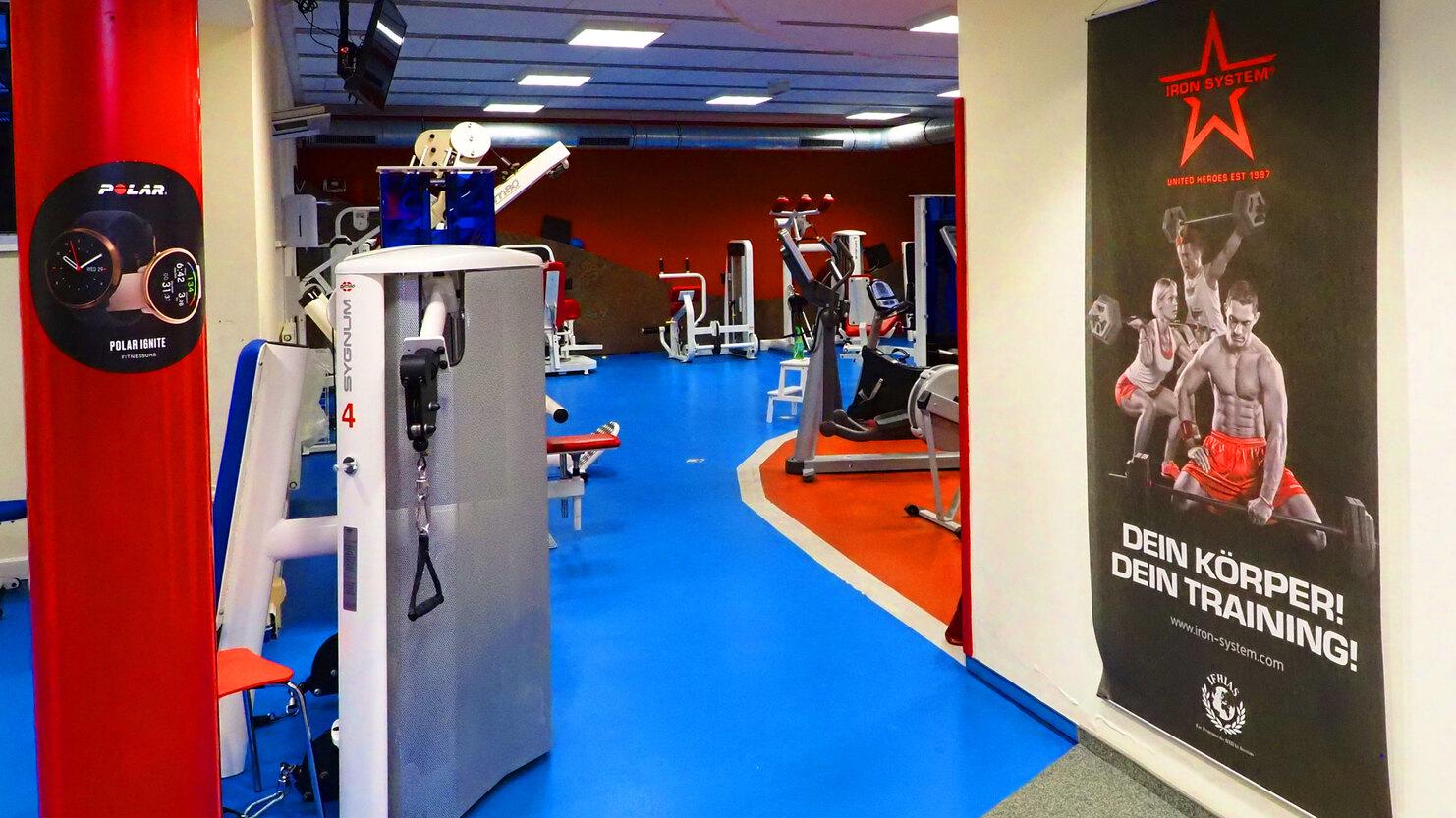 Fitnessstudio mit blauen Böden, Geräten und einem Plakat mit dem Slogan „Dein Körper! Dein Training!“.