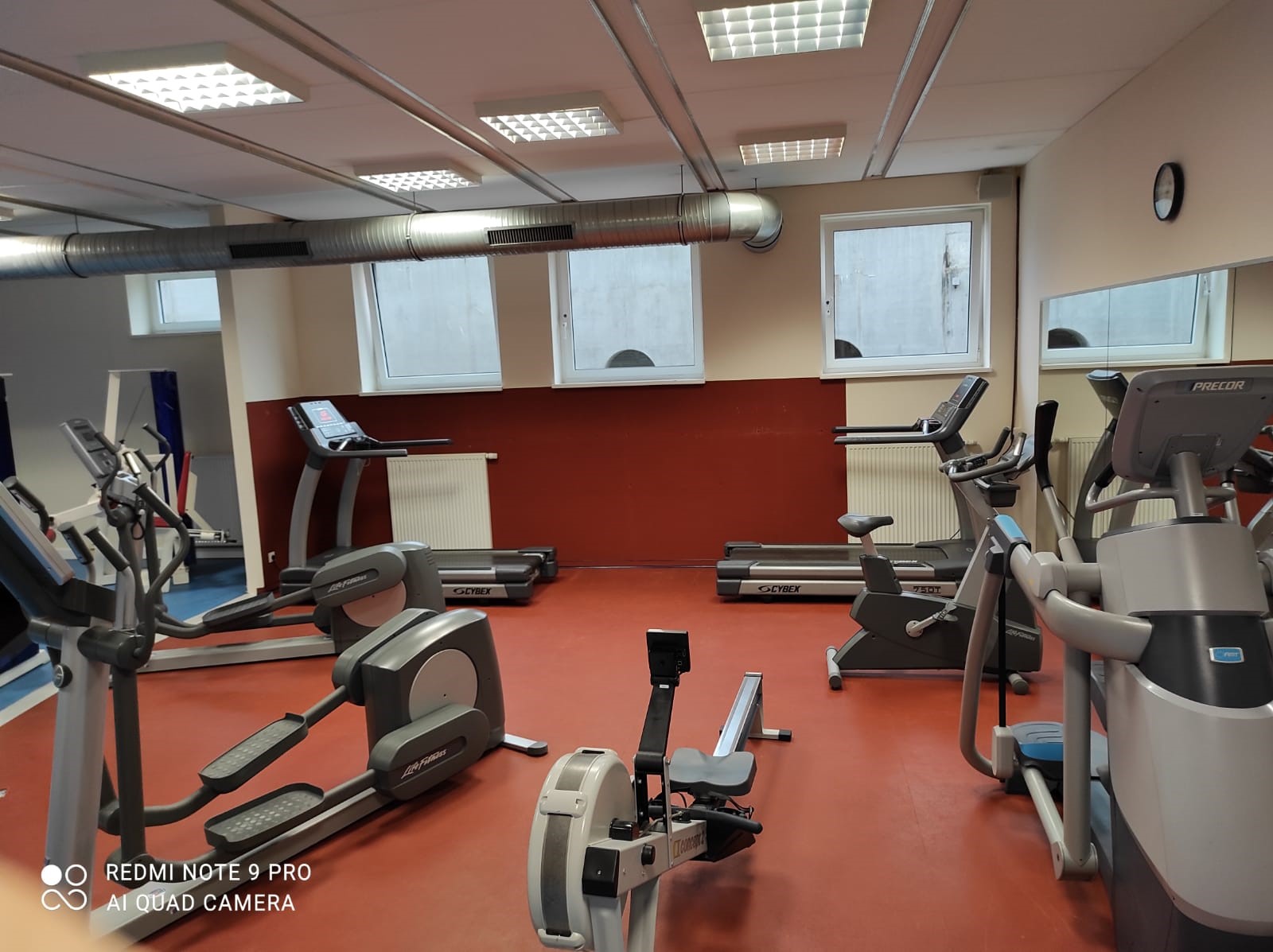 Fitnessstudio mit verschiedenen Trainingsgeräten wie Ergometern, Crosstrainern und einem Rudergerät auf rotem Boden.